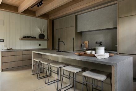 uisine contemporaine en bois et pierre, avec îlot en quartz gris, tabourets rembourrés et robinetterie noire mate, signée Ateliers Jacob.