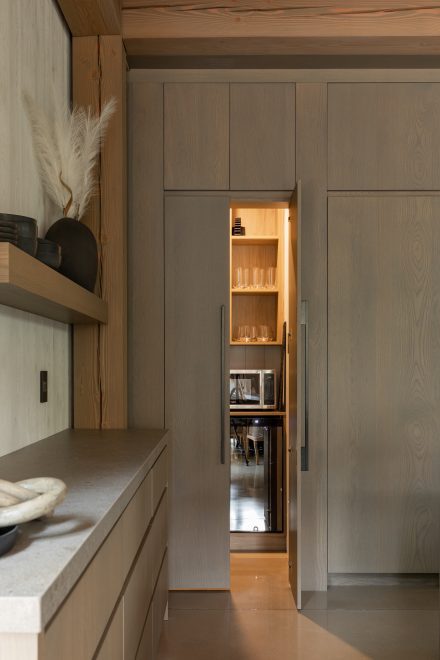 Porte de rangement dissimulée menant à un garde-manger éclairé avec micro-ondes et électroménagers intégrés, dans une cuisine haut de gamme en bois naturel.
