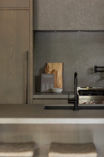  Détail d’une cuisine moderne avec surface en pierre grise, robinet noir mat, et livre Kinfolk en décoration sur fond de dosseret texturé.