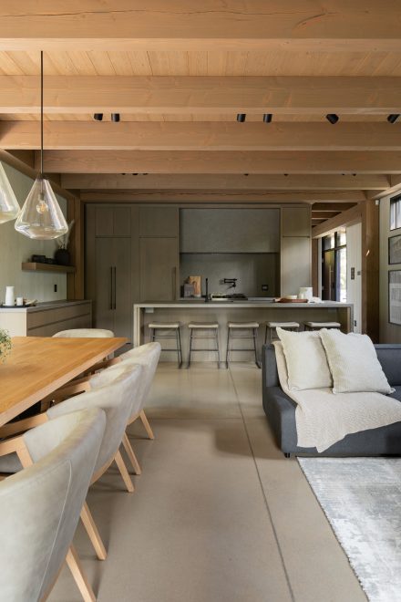 Vue sur la cuisine contemporaine à partir de la salle à manger et salon. Cuisine en bois et pierre, avec îlot en quartz gris, tabourets rembourrés et robinetterie noire mate, signée Ateliers Jacob.