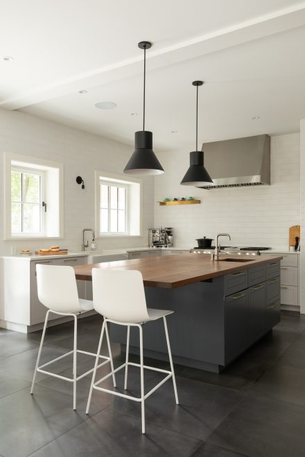 Cuisine lumineuse avec îlot central en bois, luminaires noirs suspendus et hotte professionnelle en acier inoxydable.