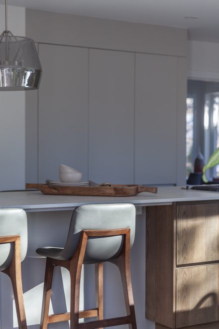 Détail d’un îlot en bois avec comptoir pâle et tabourets en cuir gris et noyer, baigné de lumière naturelle.