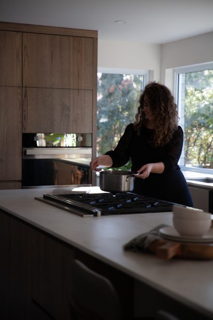 emme en pleine cuisson sur une cuisinière à gaz, dans une cuisine moderne avec armoires en bois et grandes fenêtres.