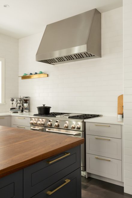 Cuisinière professionnelle en acier inoxydable avec hotte surdimensionnée, dosseret en céramique blanche et comptoir en quartz, accentuée par une étagère dorée minimaliste.