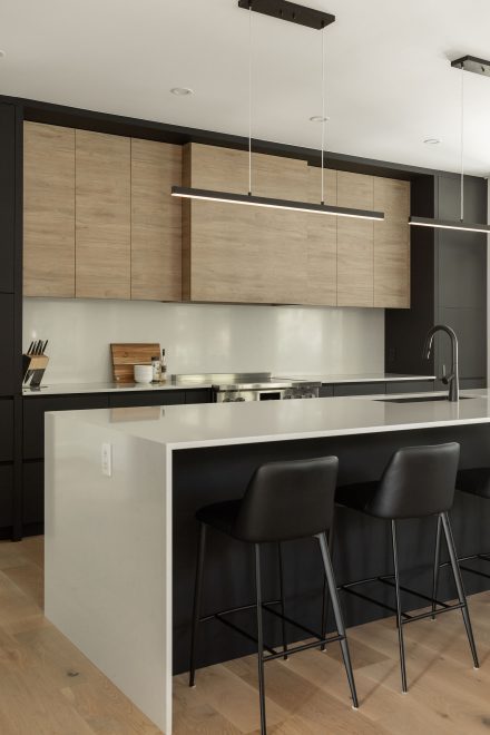 Cuisine moderne conçue par Ateliers Jacob avec armoires en bois clair et noir mat, grand îlot blanc aux lignes droites et éclairage suspendu minimaliste.