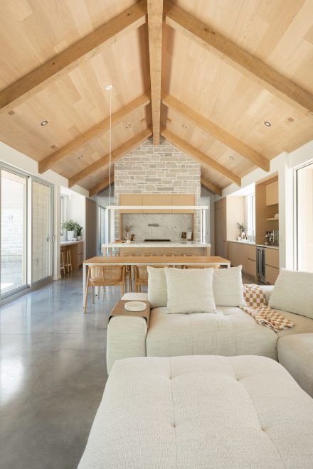 Pièce à aire ouverte avec plafond cathédrale en bois blond, cuisine en chêne clair, mur en pierre naturelle et comptoirs en marbre, combinée à un salon lumineux et mobilier texturé.