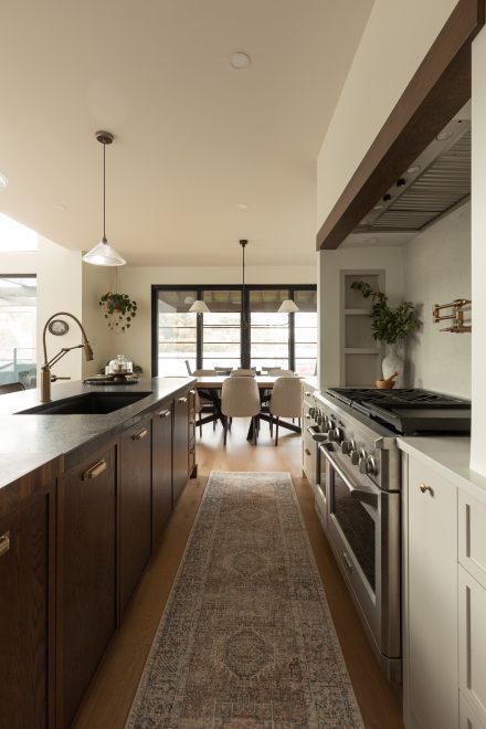 Vue traversante d’une cuisine haut de gamme avec îlot en bois foncé, tapis corridor, électroménagers professionnels et coin repas ouvert sur la terrasse.
