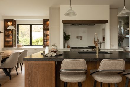 Cuisine haut de gamme avec îlot central en bois foncé, comptoir en granite foncé, coin-repas intégré et vitrines rétroéclairées de chaque côté d’une grande fenêtre.