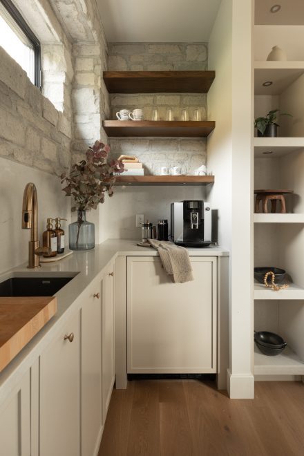 Espace café raffiné avec mur en pierre naturelle, étagères en bois massif, robinet doré et électroménagers intégrés dans une cuisine sur mesure signée Ateliers Jacob.