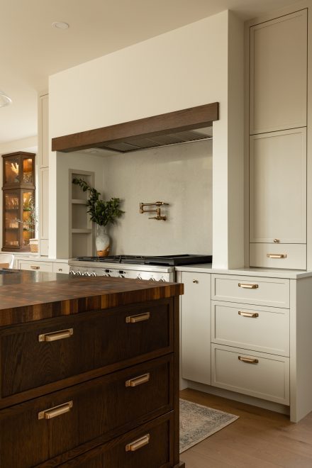 Détail du coin cuisson avec dosseret clair, robinet pot filler en laiton, hotte encastrée sous un caisson en bois et mobilier sur mesure alliant chêne foncé et teintes ivoire.