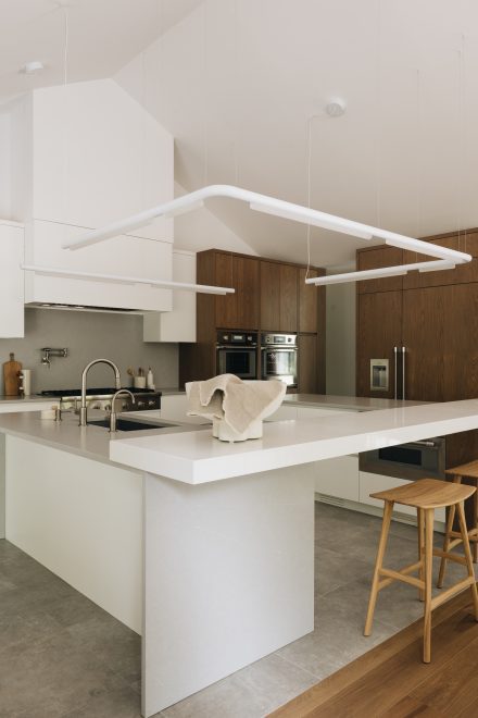 Cuisine contemporaine avec un grand îlot en cascade en pierre naturelle blanche, éclairage suspendu minimaliste, électroménagers intégrés et combinaison d’armoires en bois riche et blanc mat pour un contraste moderne et raffiné.