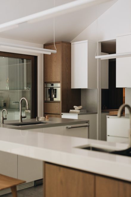 Cuisine contemporaine avec un grand îlot en cascade en pierre naturelle blanche, éclairage suspendu minimaliste, électroménagers intégrés et combinaison d’armoires en bois riche et blanc mat pour un contraste moderne et raffiné.