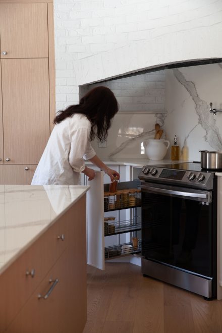 Femme ouvrant un rangement coulissant près du four, organisé avec des pots d'épices en verre, dans une cuisine sur mesure au design moderne et chaleureux.
