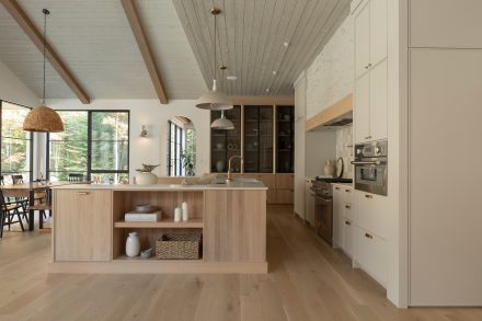 Une cuisine au design chaleureux et naturel, mettant en avant un îlot en bois clair avec des détails en laiton et un plan de travail en pierre claire. Des étagères ouvertes accueillent des paniers tressés et de la vaisselle minimaliste. Le dosseret en carreaux de céramique brillants et le mur en pierre texturée ajoutent du caractère à l’espace, tandis que deux suspensions beiges apportent une touche d’élégance discrète.