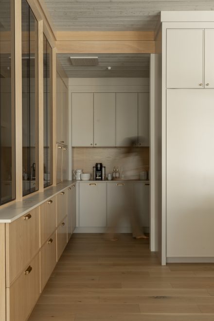 Yn garde-manger dissimulé, aux teintes douces et naturelles. Les armoires crème avec des poignées en laiton se fondent harmonieusement avec un comptoir en bois et un plancher en chêne clair. Une cloison vitrée avec des montants en bois sépare discrètement l’espace, créant une sensation d’ouverture. Une personne floue en mouvement apporte une touche de vie et de dynamisme à l’ambiance feutrée du lieu.