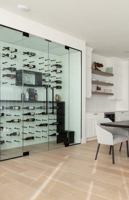 Un coin bar sophistiqué comprenant des armoires blanches, des étagères ouvertes en bois et un réfrigérateur encastré pour boissons. La cave à vin vitrée adjacente met en valeur une collection de bouteilles avec un système de rangement suspendu, apportant une touche haut de gamme à la pièce.