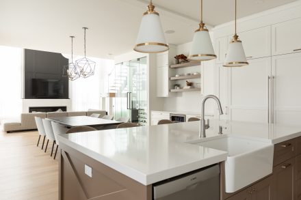 Une cuisine contemporaine avec un grand îlot en quartz blanc, accompagné de tabourets en velours beige aux pieds dorés. Les luminaires suspendus apportent une élégance intemporelle, tandis que les armoires blanches encadrent l’espace avec raffinement.