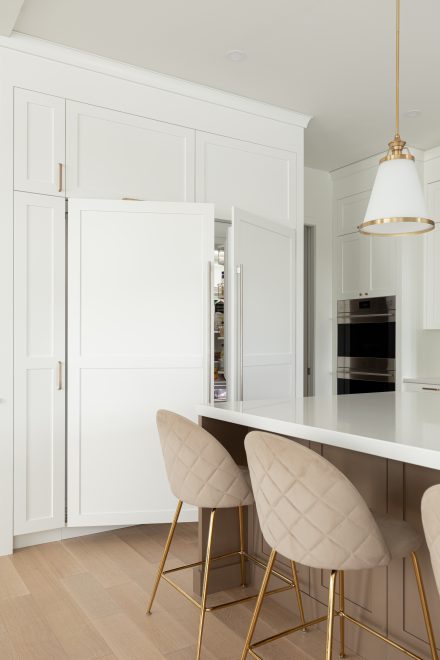 Une cuisine lumineuse et moderne avec des armoires blanches sur mesure, un îlot central en tons neutres et des accents dorés. En arrière-plan, un réfrigirateur encastré.