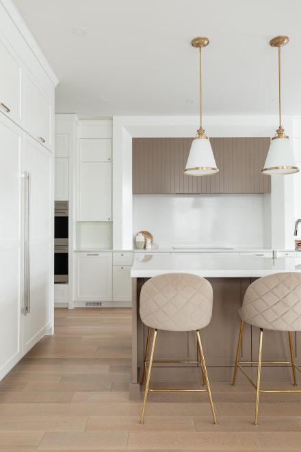 Cuisine lumineuse avec îlot taupe, tabourets à dossier matelassé et luminaires dorés, sur plancher de bois pâle.