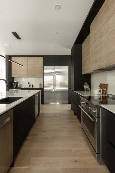 Cuisine moderne aux lignes épurées, mêlant bois clair et noir mat. L’îlot central en pierre blanche contraste avec les armoires sombres, créant un équilibre sophistiqué. Deux suspensions minimalistes illuminent l’espace.