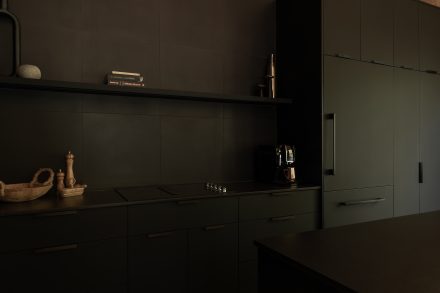 Cuisine contemporaine entièrement noire avec armoires mates, îlot central minimaliste et suspensions en fibres naturelles, contrastant avec le plafond en bois clair pour une ambiance à la fois chaleureuse et graphique.