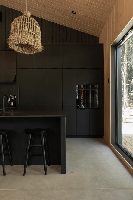Cuisine moderne entièrement noire, avec un îlot fonctionnel et des suspensions en fibres naturelles qui contrastent avec l’esthétique épurée. Un mur en bois clair et une grande fenêtre laissent entrer la lumière naturelle.

