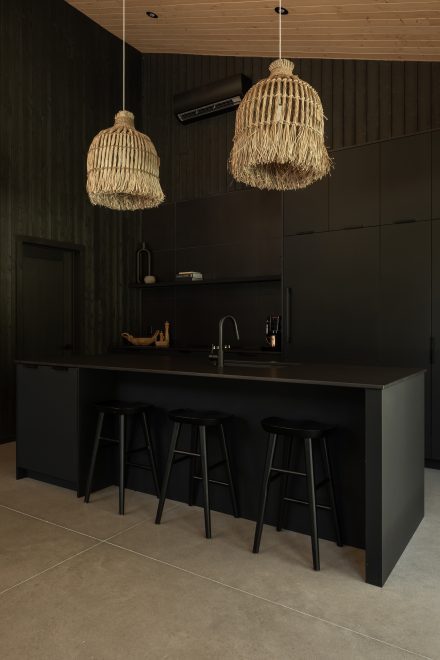 Cuisine moderne entièrement noire, avec un îlot fonctionnel et des suspensions en fibres naturelles qui contrastent avec l’esthétique épurée. Un mur en bois clair et une grande fenêtre laissent entrer la lumière naturelle.