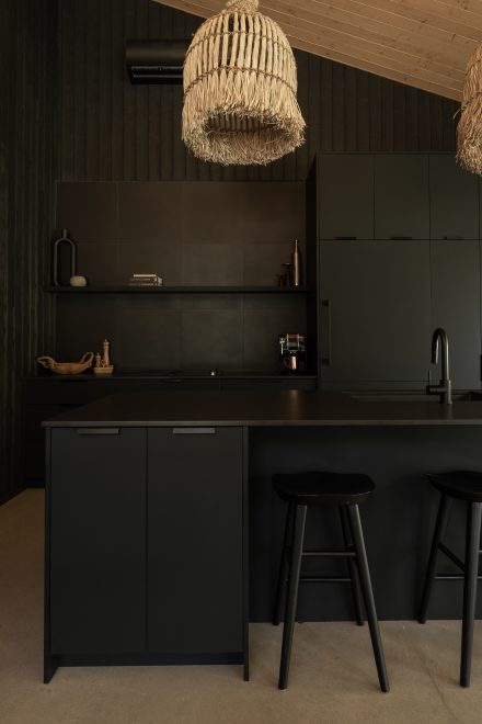 Cuisine contemporaine entièrement noire avec armoires mates, îlot central minimaliste et suspensions en fibres naturelles, contrastant avec le plafond en bois clair pour une ambiance à la fois chaleureuse et graphique.