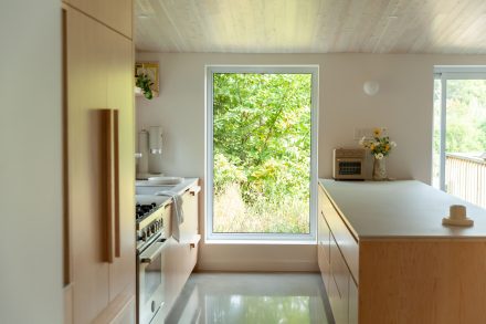 Cuisine lumineuse au design minimaliste, avec des armoires en bois pâle, un évier de style farmhouse blanc et une cuisinière à gaz, mise en valeur par une grande fenêtre offrant une vue sur la verdure extérieure.