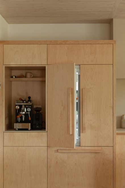 Built-in maple cabinetry featuring a concealed fridge and an integrated coffee station, showcasing minimalist design with soft wood grain and custom handles.