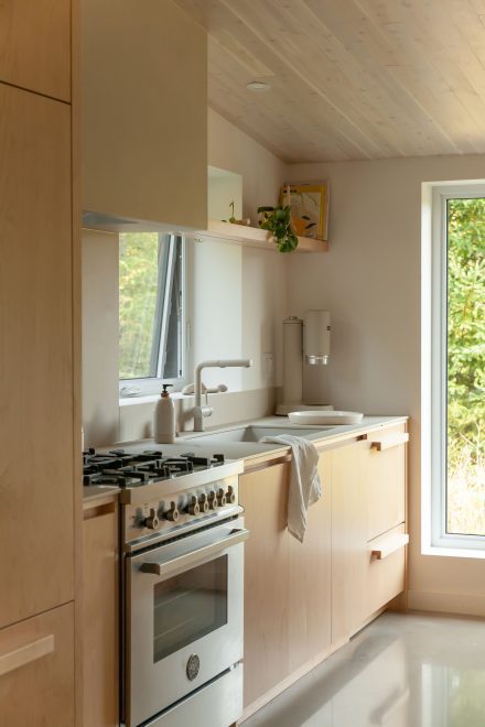 Cuisine lumineuse au design minimaliste, avec des armoires en bois pâle, un évier de style farmhouse blanc et une cuisinière à gaz, mise en valeur par une grande fenêtre offrant une vue sur la verdure extérieure.