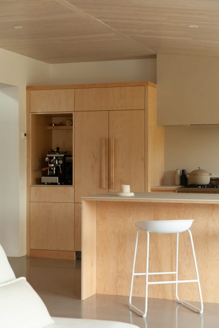 Cuisine lumineuse au design minimaliste, avec des armoires en bois pâle, un évier de style farmhouse blanc et une cuisinière à gaz, mise en valeur par une grande fenêtre offrant une vue sur la verdure extérieure.