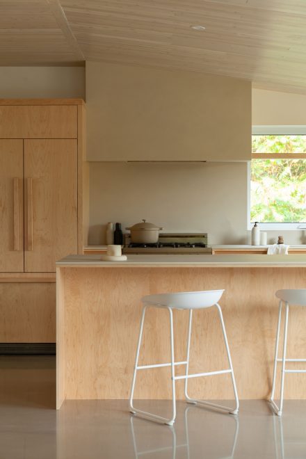 Cuisine lumineuse et épurée avec un îlot central en bois clair et un comptoir en pierre beige. Deux tabourets minimalistes blancs contrastent avec les matériaux naturels, offrant un espace de repas moderne et chaleureux.