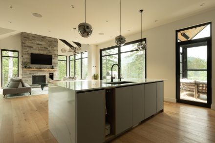 Îlot de cuisine en quartz avec évier intégré sur fond de salon lumineux, accentué par un foyer en pierre naturelle, de grandes fenêtres noires et une vue sur la forêt.