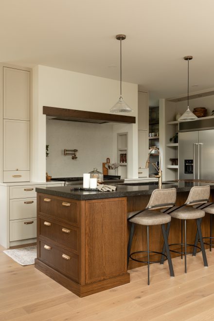 Vue d’ensemble d’une cuisine chaleureuse avec îlot en bois foncé, tabourets en cuir beige et luminaires suspendus en verre texturé.