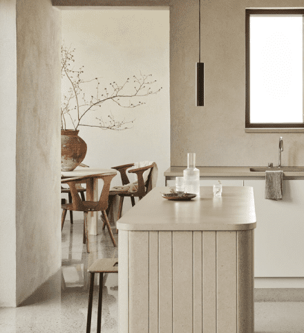 Cuisine minimaliste avec îlot en béton beige texturé, comptoir arrondi et tabouret en bois. En arrière-plan, salle à manger avec table ronde et chaises sculpturales en bois foncé, mise en valeur par un grand vase rustique avec branches séchées. L’ensemble baigne dans une lumière douce, soulignant les textures naturelles et l’esthétique épurée du lieu.