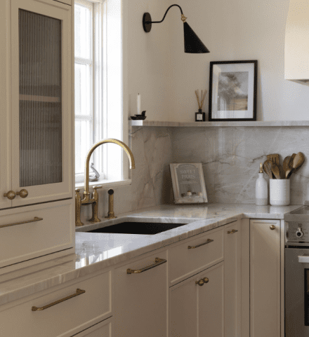 Cuisine Ateliers Jacob avec évier intégré dans un comptoir en pierre pâle, armoires crème, robinetterie dorée et dosseret assorti.