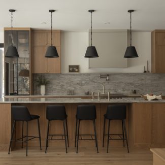 Cuisine contemporaine sur mesure conçue par Ateliers Jacob, combinant armoires en chêne naturel, quartz gris et hotte intégrée discrète. L’îlot central en pierre naturelle offre des assises noires modernes et un robinet doré, soulignant le design haut de gamme et l’élégance fonctionnelle.