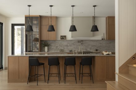Cuisine contemporaine sur mesure conçue par Ateliers Jacob, combinant armoires en chêne naturel, quartz gris et hotte intégrée discrète. L’îlot central en pierre naturelle offre des assises noires modernes et un robinet doré, soulignant le design haut de gamme et l’élégance fonctionnelle.