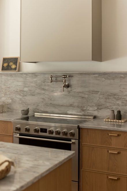 Cuisine contemporaine sur mesure conçue par Ateliers Jacob, combinant armoires en chêne naturel, quartz gris et hotte intégrée discrète. L’îlot central en pierre naturelle offre des assises noires modernes et un robinet doré, soulignant le design haut de gamme et l’élégance fonctionnelle.