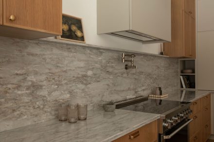 Cuisine contemporaine sur mesure conçue par Ateliers Jacob, combinant armoires en chêne naturel, quartz gris et hotte intégrée discrète. L’îlot central en pierre naturelle offre des assises noires modernes et un robinet doré, soulignant le design haut de gamme et l’élégance fonctionnelle.