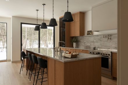 Cuisine contemporaine sur mesure conçue par Ateliers Jacob, combinant armoires en chêne naturel, quartz gris et hotte intégrée discrète. L’îlot central en pierre naturelle offre des assises noires modernes et un robinet doré, soulignant le design haut de gamme et l’élégance fonctionnelle.