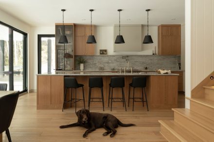 Cuisine contemporaine sur mesure conçue par Ateliers Jacob, combinant armoires en chêne naturel, quartz gris et hotte intégrée discrète. L’îlot central en pierre naturelle offre des assises noires modernes et un robinet doré, soulignant le design haut de gamme et l’élégance fonctionnelle.