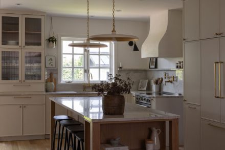 Cuisine haut de gamme conçue par Ateliers Jacob à Montréal, mariant des armoires crème avec des poignées en laiton, un îlot en bois clair et un comptoir en marbre. La hotte sculpturale et les luminaires suspendus ajoutent une touche artisanale et contemporaine à l’ensemble.