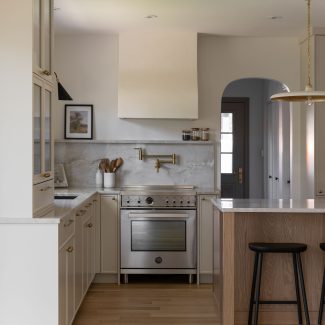 Cuisine haut de gamme conçue par Ateliers Jacob à Montréal, mariant des armoires crème avec des poignées en laiton, un îlot en bois clair et un comptoir en marbre. La hotte sculpturale et les luminaires suspendus ajoutent une touche artisanale et contemporaine à l’ensemble.