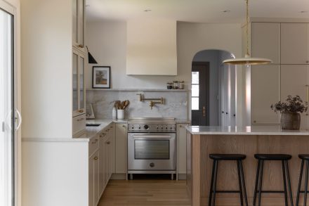 Cuisine haut de gamme conçue par Ateliers Jacob à Montréal, mariant des armoires crème avec des poignées en laiton, un îlot en bois clair et un comptoir en marbre. La hotte sculpturale et les luminaires suspendus ajoutent une touche artisanale et contemporaine à l’ensemble.