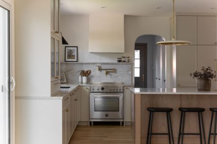 Cuisine haut de gamme conçue par Ateliers Jacob à Montréal, mariant des armoires crème avec des poignées en laiton, un îlot en bois clair et un comptoir en marbre. La hotte sculpturale et les luminaires suspendus ajoutent une touche artisanale et contemporaine à l’ensemble.