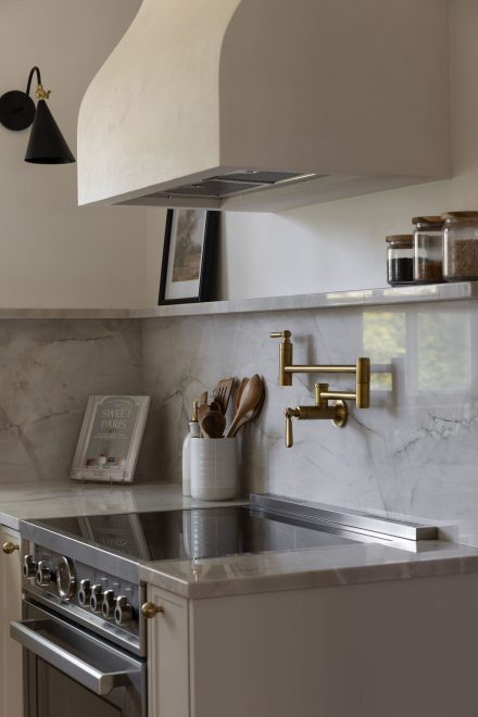 Cuisine haut de gamme conçue par Ateliers Jacob à Montréal, mariant des armoires crème avec des poignées en laiton, un îlot en bois clair et un comptoir en marbre. La hotte sculpturale et les luminaires suspendus ajoutent une touche artisanale et contemporaine à l’ensemble.