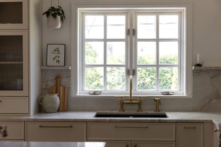 Cuisine haut de gamme conçue par Ateliers Jacob à Montréal, mariant des armoires crème avec des poignées en laiton, un îlot en bois clair et un comptoir en marbre. La hotte sculpturale et les luminaires suspendus ajoutent une touche artisanale et contemporaine à l’ensemble.