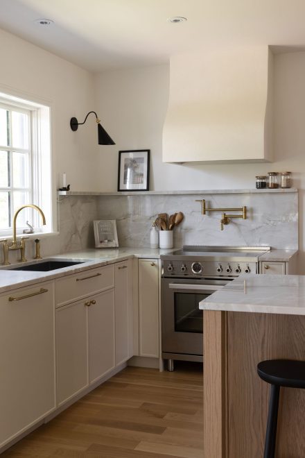 Cuisine haut de gamme conçue par Ateliers Jacob à Montréal, mariant des armoires crème avec des poignées en laiton, un îlot en bois clair et un comptoir en marbre. La hotte sculpturale et les luminaires suspendus ajoutent une touche artisanale et contemporaine à l’ensemble.